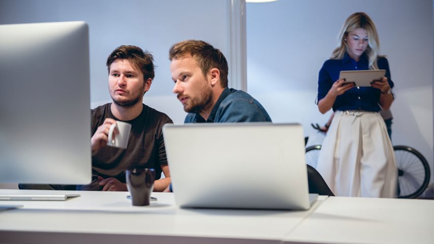 To mænd kigger på computer og en kvinde i baggrunden kigger på tablet